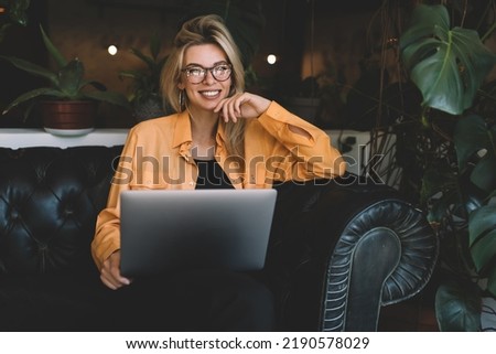Happy millennial woman in optical eyewear glasses holding digital netbook device and smiling at camera, portrait of cheerful Caucaisan female blogger with modern laptop technology posing indoors Royalty-Free Stock Photo #2190578029