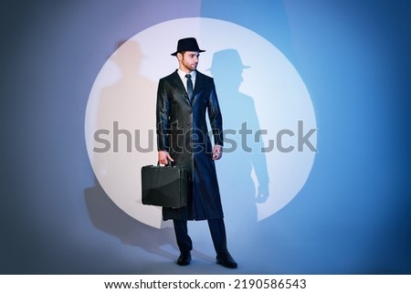 Full length portrait of handsome man in black coat and hat holding briefcase posing in the spotlight on studio background. noir film style. Private detective, spy, investigation concept.  Royalty-Free Stock Photo #2190586543