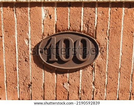 House number (address) one hundred (100) sign, rusty numbered plate (closeup) against red (orange) bricks wall of old building background. Grunge, texture, wallpaper, rust