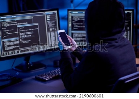 cybercrime, hacking and technology concept - hacker with smartphone using computer virus program for cyber attack in dark room Royalty-Free Stock Photo #2190600601