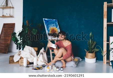 Carefree young female artist posing for smartphone camera selfie near easel and paints showing in blog her hobby. Creative smiling brunette woman photographing herself near draw oil picture 