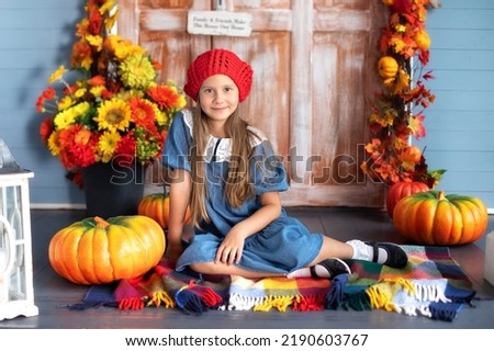 Little happy girl with pumpkin and smiling at camera while sitting on house porch and waiting to trick or treat on Halloween. Child is sitting next to pumpkins near door of house. Fall home decoration