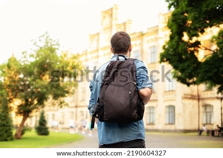 Student university standing with his back to the camera and his backpack on one shoulder and walking in university campus, education concept. Young man walking down street with a backpack. Back view.
