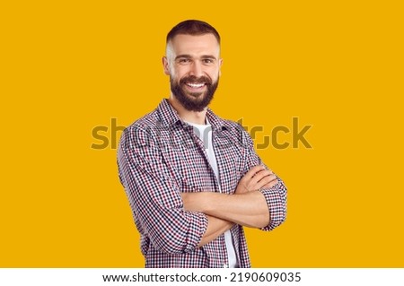 Studio shot of a happy young guy. Cheerful handsome bearded young man wearing a casual shirt standing with his arms folded isolated on a yellow color background, looking at the camera and smiling Royalty-Free Stock Photo #2190609035