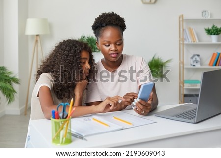 Mother helps her teenage daughter complete her school homework while using her smartphone. African American family sitting in front of notebook and laptop and looking at mobile phone screen together Royalty-Free Stock Photo #2190609043