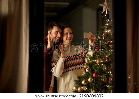 winter holidays and people concept - happy couple with smartphone taking selfie or having video call and waving hand at christmas tree at home