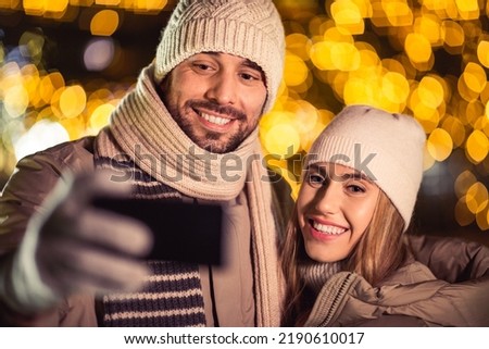winter holidays and people concept - happy smiling couple taking selfie with smartphone and hugging over christmas lights in evening
