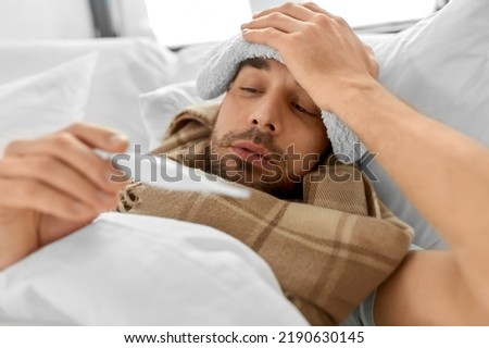 people, health and fever concept - sick man with cold compress on his forehead measuring temperature by thermometer lying in bed at home Royalty-Free Stock Photo #2190630145