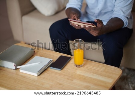 Cropped image of businessman sitting in front of coffee table with planner, smartphone and glass of orange juice and reading e-book
