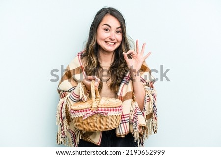 Young caucasian woman holding a picnic basket isolated on blue background cheerful and confident showing ok gesture.