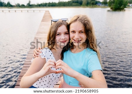 Two teens girl friends their hands fingers make heart symbol standing on a wooden bridge with lake on background. Childhood, growing up, friendship, summer camp. Royalty-Free Stock Photo #2190658905