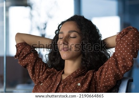 Close-up photo portrait of young beautiful curly Arab woman resting indoors at home, woman with hands behind head resting sitting on sofa near window, eyes closed breathing, meditating and dreaming Royalty-Free Stock Photo #2190664865