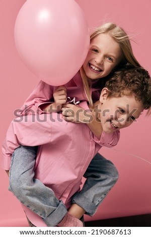 joyful, happy children of school age, on a pink background in pink clothes play and the boy rolls the girl with a balloon on her back