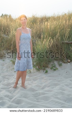 Attractive short haired woman in her thirties, dressed in light summer dress standing barefoot on the beach with grass in the background. Concept for vacation, relaxation, mental health.