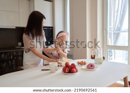 Mother and daughter preparing a sweet cake using flour, milk, sitting on chairs at a table in a modern kitchen. Girl holding a whisk, stirring eggs in a bowl, preparing pancake dough with her mom.