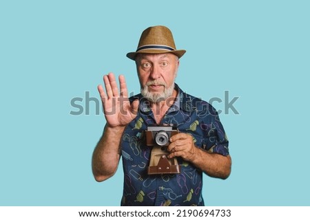 Strike a pose. Eldery man with gray beard and straw hat holding old camera rise his hand up to take picture. Blue background portrait