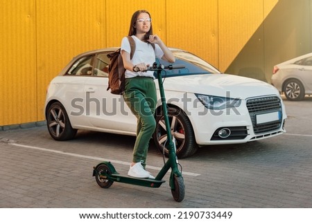 Young woman in white sneakers standing on electric scooter in front of the white car