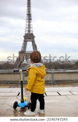 Faceless small boy in yellow coat and jeans with kick scooter stands on the Eiffel tower background in Paris