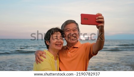 asian elderly couple standing on the beach and taking selfie happily in sunset background