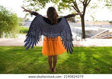 A girl dressed as a witch with an orange skirt and black wings stretches out her arms. The picture is taken from the girl's back. Trick or treat. 31st October. Happy halloween.