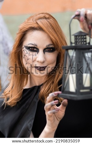 Attractive beautiful woman in a Halloween costume black cloak and scary make-up. Young girl in a look of a witch dressed up for a party making fairy potion holding black lantern. High quality photo