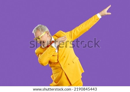 Funny happy confident handsome senior man in yellow suit and eyeglasses doing dab moves. Cheerful eccentric mature guy wearing trendy outfit dancing isolated on purple background. Fashion concept Royalty-Free Stock Photo #2190845443