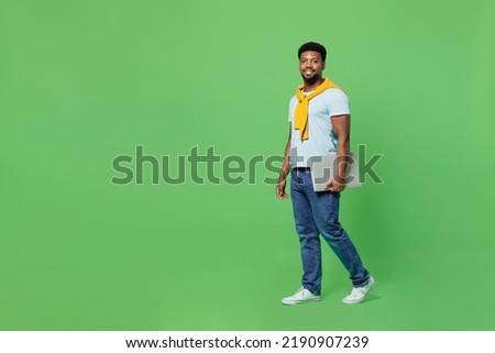 Full body young fun man of African American ethnicity 20s wear blue t-shirt hold closed laptop pc computer walking going isolated on plain green background studio portrait. People lifestyle concept