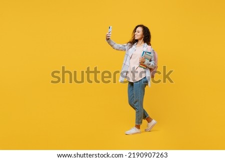 Full body young black teen girl student she wear casual clothes backpack bag hold books do selfie shot on mobile cell phone isolated on plain yellow background. High school university college concept