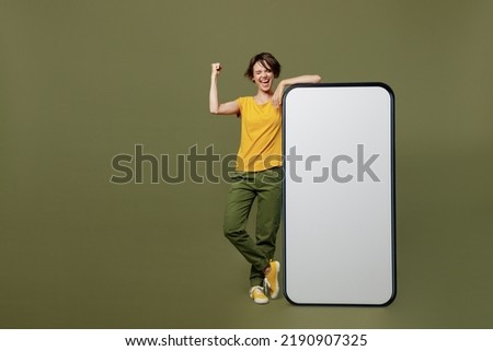 Full body young woman she 20s wear yellow t-shirt near big huge blank screen mobile cell phone with workspace copy space mockup area do winner gesture isolated on plain olive green khaki background