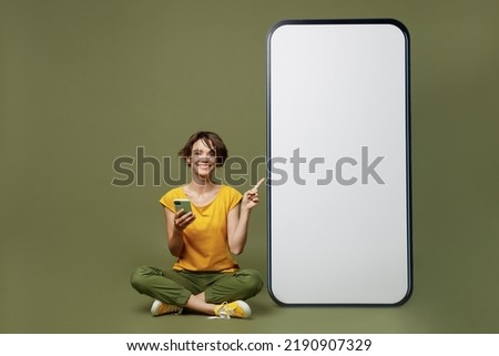 Full body young woman she 20s wear yellow t-shirt point on big huge blank screen mobile cell phone with workspace copy space mockup area use smartphone isolated on plain olive green khaki background