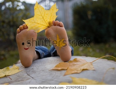 yellow autumn leaf between the toes on the bare feet of a toddler child. painted smile on the feet. indulge, positive thinking, happy childhood. Hello, Autumn. seasonal fun photo ideas