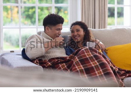 Happy biracial couple sitting on couch under blanket, drinking coffee and laughing in living room. Inclusivity, domestic life, leisure time and togetherness concept. Royalty-Free Stock Photo #2190909141