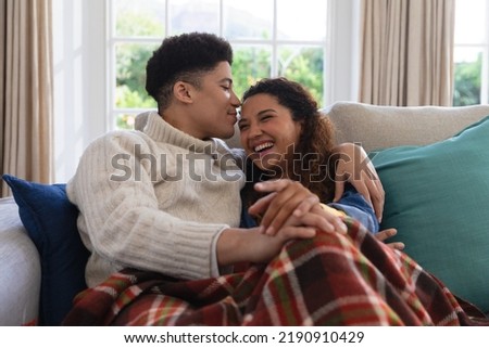 Happy biracial couple sitting on couch under blanket, laughing and embracing in living room. Inclusivity, domestic life, leisure time and togetherness concept. Royalty-Free Stock Photo #2190910429