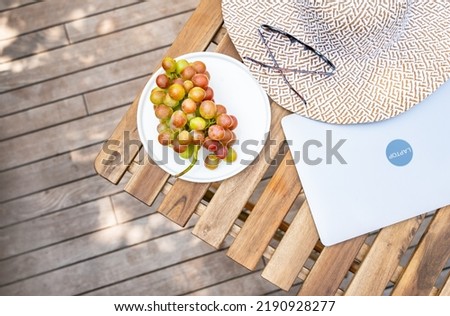 Closed laptop, grape, stylish hat, sunglasses are on a wooden table on a summer terrace with copy space. Summer mood, vacation, freelance, distance work concept. Top view. Background with copy space.