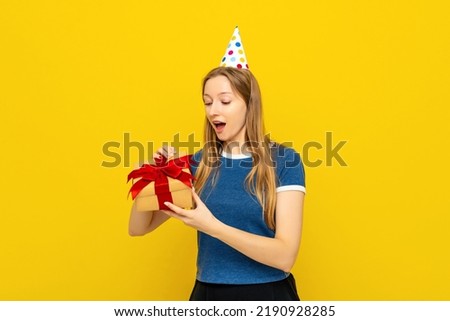 Smiling pretty girl in a birthday cap and blue t shirt opening gift box and smiling happy. Holidays and birthday concept. Colorful studio portrait with yellow background