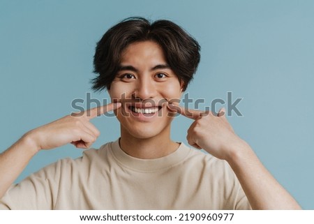 Young handsome asian boy stretching his smile with fingers and looking at camera while standing over isolated blue background
