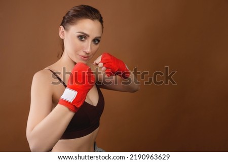 A young woman in boxing gloves poses in a fighting stance and looks at the camera. Attractive female boxer ready to fight on brown background