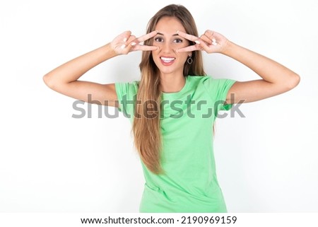 Cheerful positive young beautiful woman wearing green T-shirt over white background shows v-sign near eyes open mouth