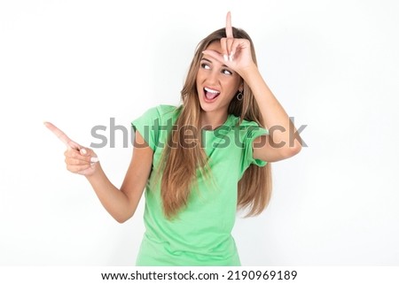 young beautiful woman wearing green T-shirt over white background showing loser sign and pointing at empty space