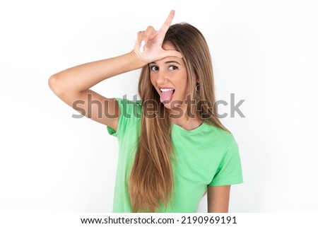 Funny young beautiful woman wearing green T-shirt over white background makes loser gesture mocking at someone sticks out tongue making grimace face.