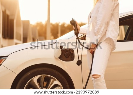 Holding charger. Young woman in white clothes is with her electric car at daytime.