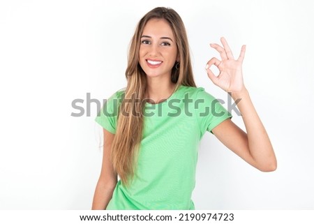young beautiful woman wearing green T-shirt over white background hold hand arm okey symbol toothy approve advising novelty news