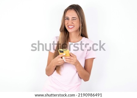 Pleased young beautiful woman wearing pink T-shirt over white background using self phone and looking and winking at the camera. Flirt and coquettish concept.
