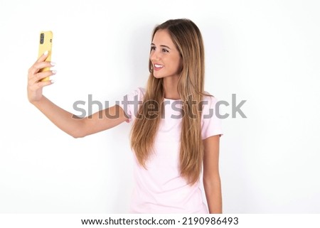 Isolated shot of pleased cheerful young beautiful woman wearing pink T-shirt over white background, makes selfie with mobile phone. People, technology and leisure concept