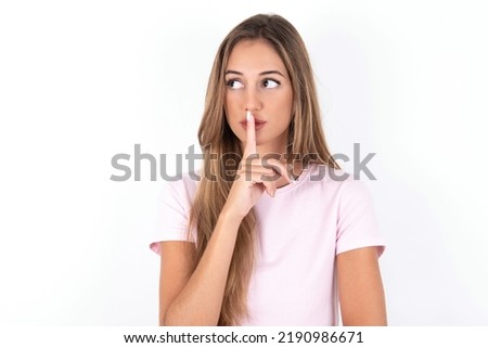 young beautiful woman wearing pink T-shirt over white background makes silence gesture, keeps index finger to lips makes hush sign. Asks not to share secret.