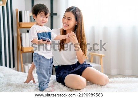 Little Asian boy hold mobile phone and stand on bed also smile with happiness to stay with his mother sit beside in bedroom with day light.