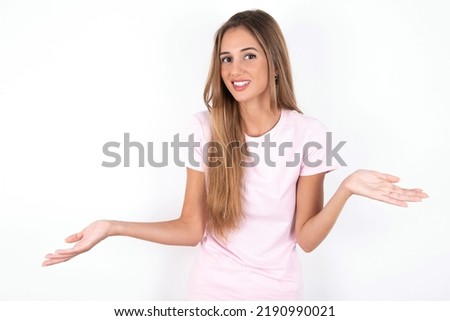 So what? Portrait of arrogant young beautiful woman wearing pink T-shirt over white background shrugging hands sideways smiling gasping indifferent, telling something obvious. Royalty-Free Stock Photo #2190990021