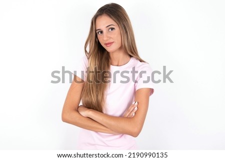 Waist up shot of  self confident young beautiful woman wearing pink T-shirt over white background has broad smile, crosses arms, happy to meet with colleagues.