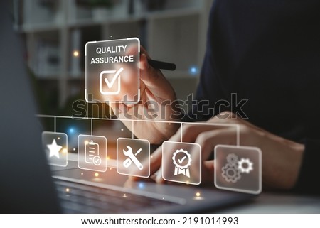 Hand of businessman holding a pen pointing to Quality Assurance icon for service guarantee on laptop screen,  Standards control and certification concept.