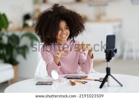 Smiling adorable black school girl famous vlogger steraming from home, sitting at kitchen table, showing her followers kids cosmetics, recording video on cell phone, copy space